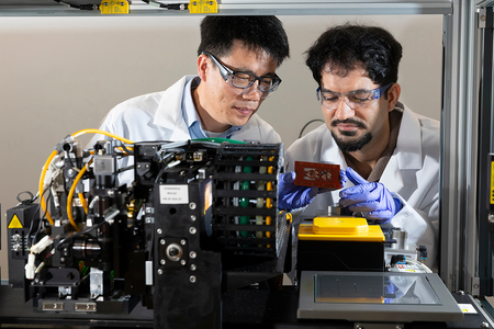 Two researchers wearing lab coats, safety glasses, and gloves examine a laboratory manufacturing machine with electronic equipment and a circuit sample.