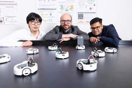 Three researchers lean over a table surrounded by multiple small robots during a laboratory demonstration with research posters in the background.