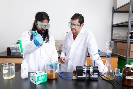 Researcher in safety goggles uses a pipette to transfer liquid into a beaker containing a cloudy brown sample while a colleague observes beside lab equipment.