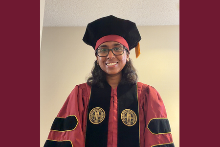 woman in garnet and black phd hood cap gown and black glasses smiling