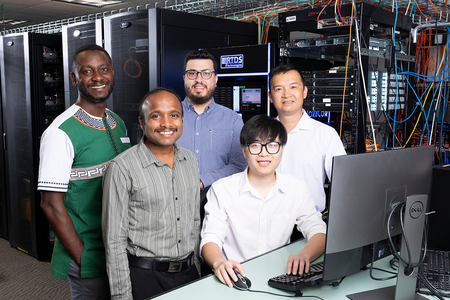 Five researchers stand together in a server room beside a desktop computer, with server racks, networking equipment, and cabling visible in the background.