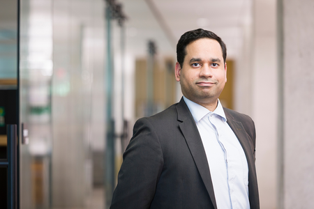 A faculty member wearing a dark business suit and light dress shirt stands in a hallway with glass-walled offices in the background.