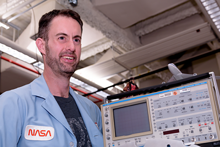 man in blue NASA shirt and beard smiling looking to right past camera