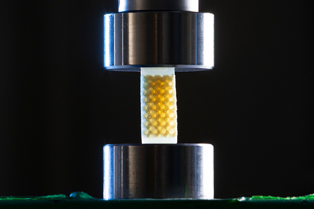 Close-up of a small lattice foam sample with a repeating internal structure positioned between two cylindrical metal plates of a compression testing machine.