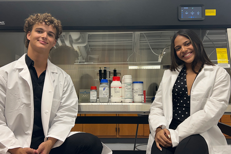 Two students in white lab coats sit on either side of a biosafety cabinet in a lab. Inside the cabinet, pipettes hang above lab supply containers, including bottles hand-labeled “PelliTect.”