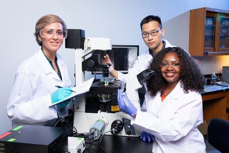 Three researchers wearing white lab coats and protective eyewear pose around a laboratory microscope. A woman on the left holds a notebook and pen, a man behind the microscope adjusts the equipment, and a woman on the right smiles while holding a clear sample tube.