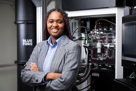 A smiling woman with long burgundy braids stands with her arms crossed in front of laboratory equipment, wearing a gray pinstripe blazer over a light blue shirt. Metal pipes, valves and a large black Bluefors cylindrical unit are visible behind her.
