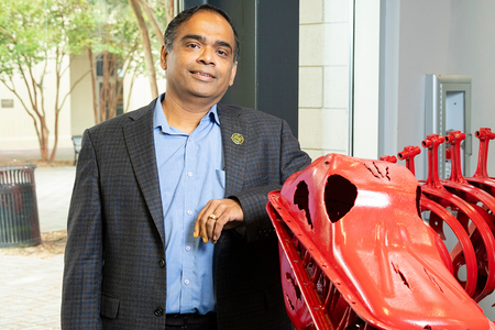 A man in a gray plaid blazer and light blue shirt stands smiling with one arm resting on a red metal sculpture of a dinosaur skeleton inside a building lobby, with a brick courtyard visible through the windows behind him.