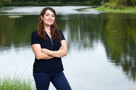 Woman in a navy blouse standing with arms crossed in front of a calm lake, smiling at the camera.