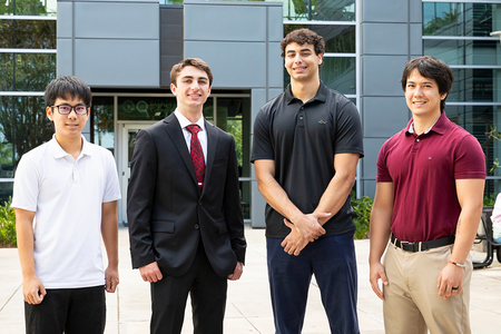 Four young men pose together in an outdoor courtyard in front of a modern glass-and-metal building.