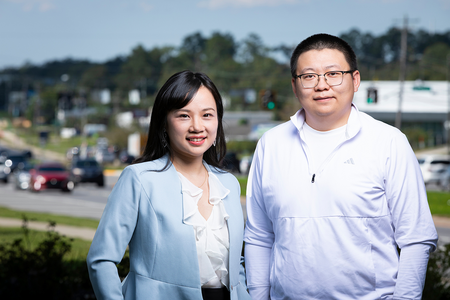 A woman with long black hair wearing a light blue blazer stands beside a man with short black hair, glasses, and a white quarter-zip pullover. Both smile at the camera, with passing vehicles and trees blurred in the background.