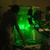 Two researchers adjust a metal-framed experimental rig in a dark lab illuminated by bright green laser light, with visible beams and cables surrounding the apparatus.
