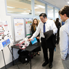A student in a white shirt and tie demonstrates a cylindrical engineering device with attached wiring on a table while two attendees observe and ask questions, with a project poster on buoyancy control displayed beside them in a hallway setting.