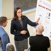 Presenter points to a section of a research poster while explaining findings to two individuals standing nearby.