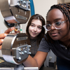 Two women closely observing and adjusting a materials testing device, with one reaching toward the sample mounted between metal grips while the other looks on.