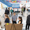 Overhead view of multiple groups of people discussing research posters arranged on easels in a bright atrium space.