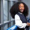 A young woman with curly hair smiles while leaning against a blue railing, wearing a navy and white jacket with Boeing TMFC Scholar Program text on the sleeve.