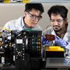 Two researchers wearing lab coats, safety glasses, and gloves examine a laboratory manufacturing machine with electronic equipment and a circuit sample.