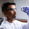 A researcher wearing a lab coat, safety glasses, and a purple nitrile glove examines a small cylindrical sample while working in a laboratory.