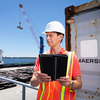 Man wearing a white hard hat and orange reflective safety vest holds an open black tablet while standing beside a shipping container at a waterfront cargo facility, with a ship and crane in the background.