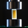 Close-up of a small lattice foam sample with a repeating internal structure positioned between two cylindrical metal plates of a compression testing machine.