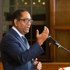 man in blue suit and black glasses talking at podium with hand guestures