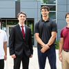Four young men pose together in an outdoor courtyard in front of a modern glass-and-metal building.