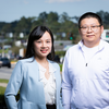 A woman with long black hair wearing a light blue blazer stands beside a man with short black hair, glasses, and a white quarter-zip pullover. Both smile at the camera, with passing vehicles and trees blurred in the background.