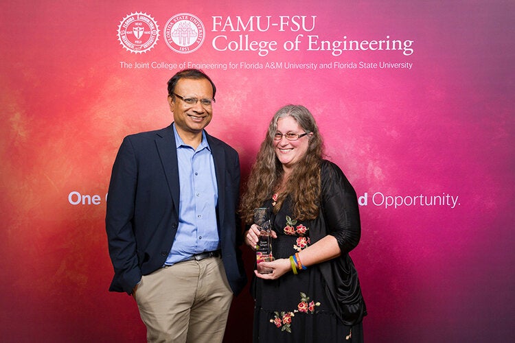 Two individuals pose in front of a FAMU-FSU College of Engineering backdrop, with the person on the right holding a clear glass award and smiling at the camera.