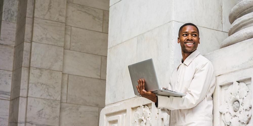 Black male holding a laptop while smiling with his head turned to the left.